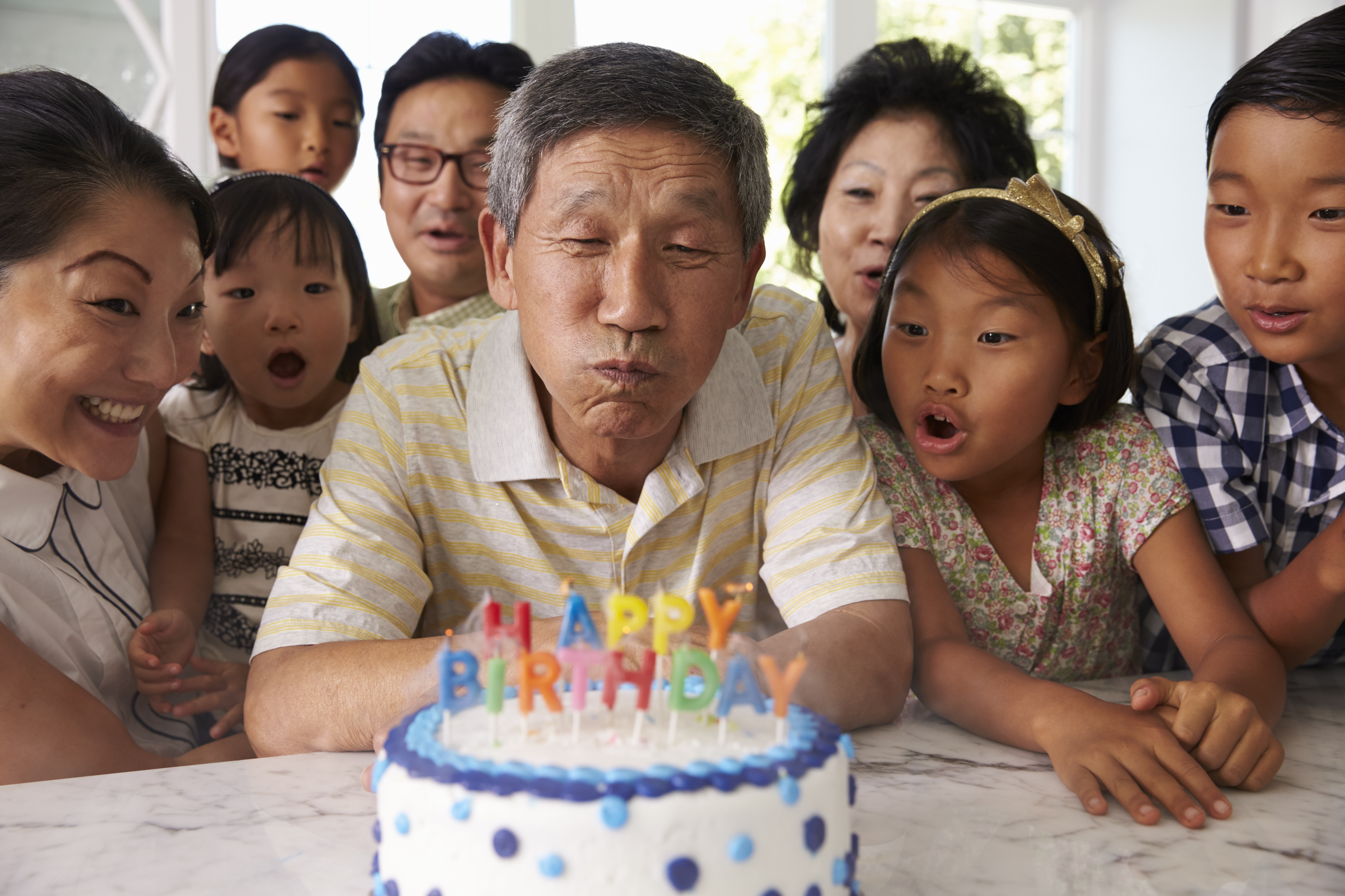  Familia de abuelos, padres y nietos rodeando un pastel y soplando las velas que dicen "Feliz cumpleaños".​​ 