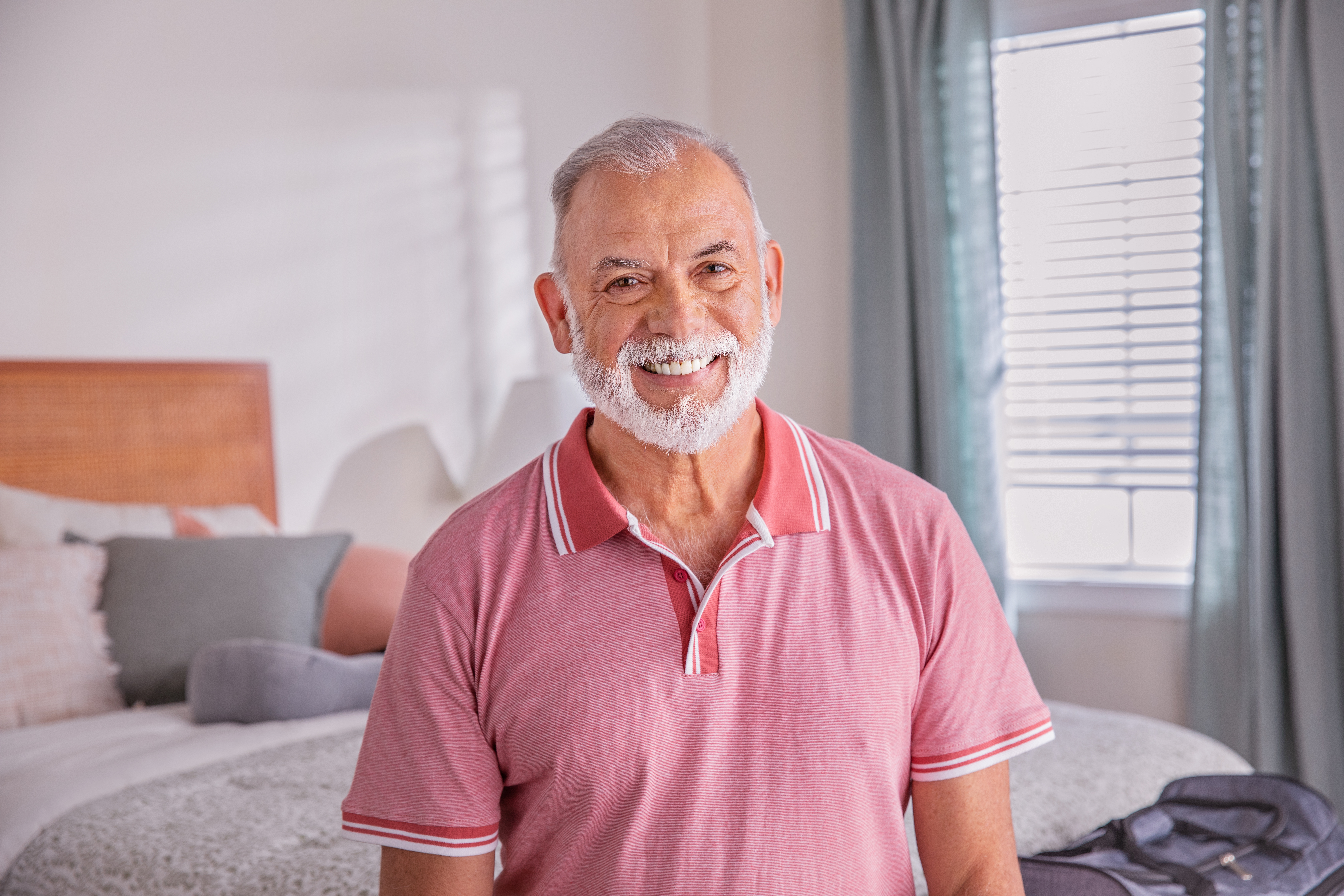 Senior man poses in bedroom sitting on bed next to suitcase​​ 