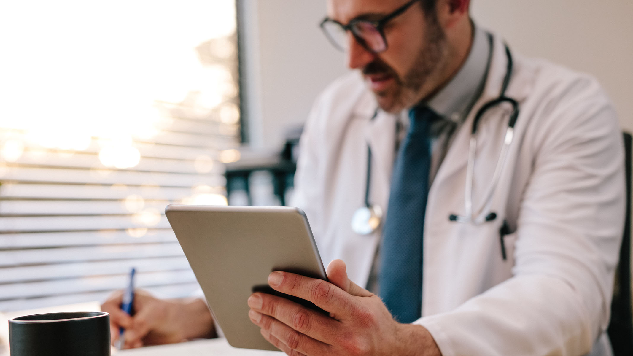 Un profesional del cuidado de la salud usando su tablet y tomando notas en su escritorio.​​ 
