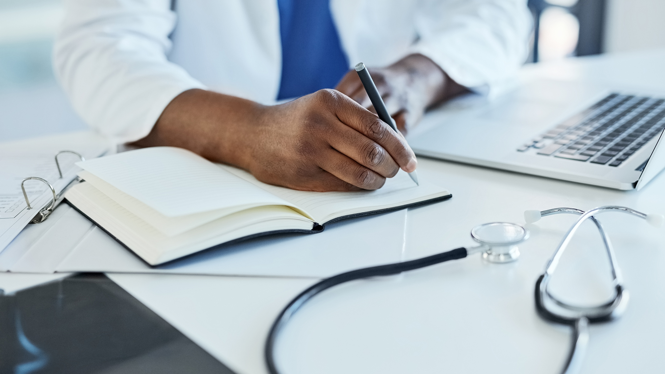 Primer plano de un médico usando un computador portátil y tomando notas.​​ 