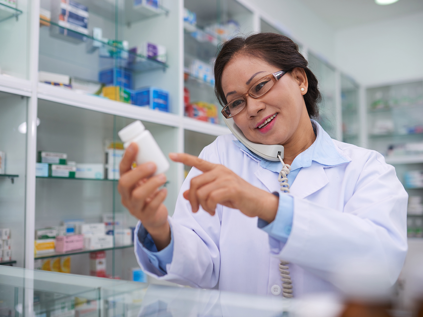 Farmacéutico leyendo la etiqueta de un medicamento mientras habla por teléfono.​​ 