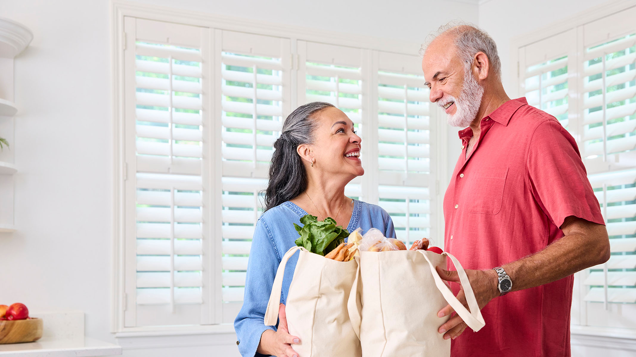 Pareja de personas mayores con bolsas de comestibles en la cocina.​​ 