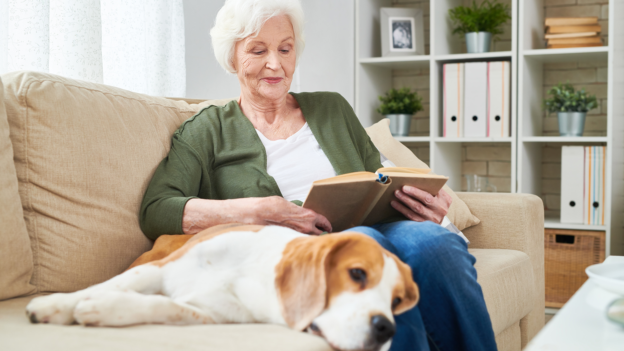 Mujer mayor leyendo un libro en el sofá junto a un perro.​​ 