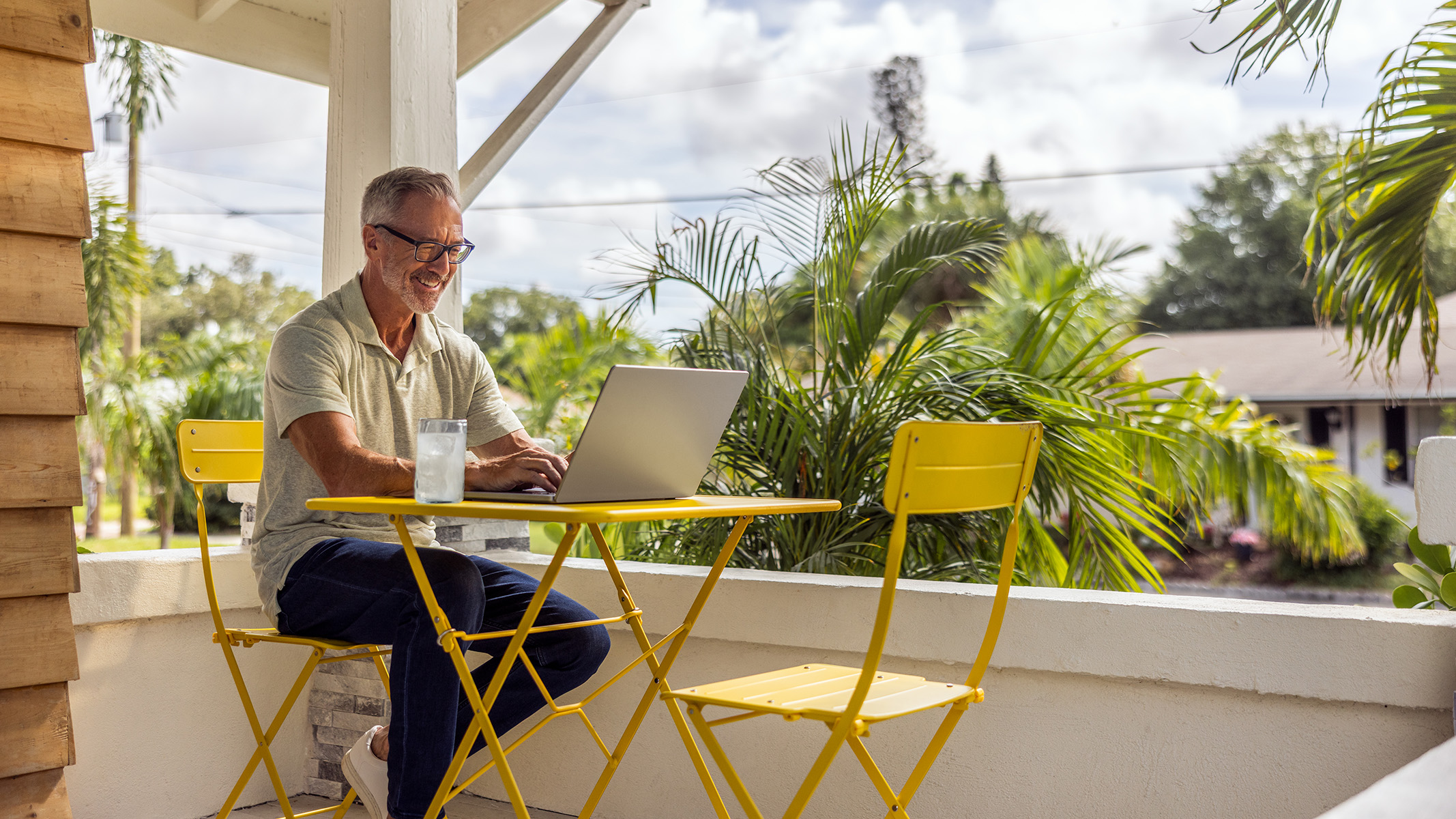 Una persona mayor sentada al aire libre en un entorno tropical utilizando su computador portátil.​​ 