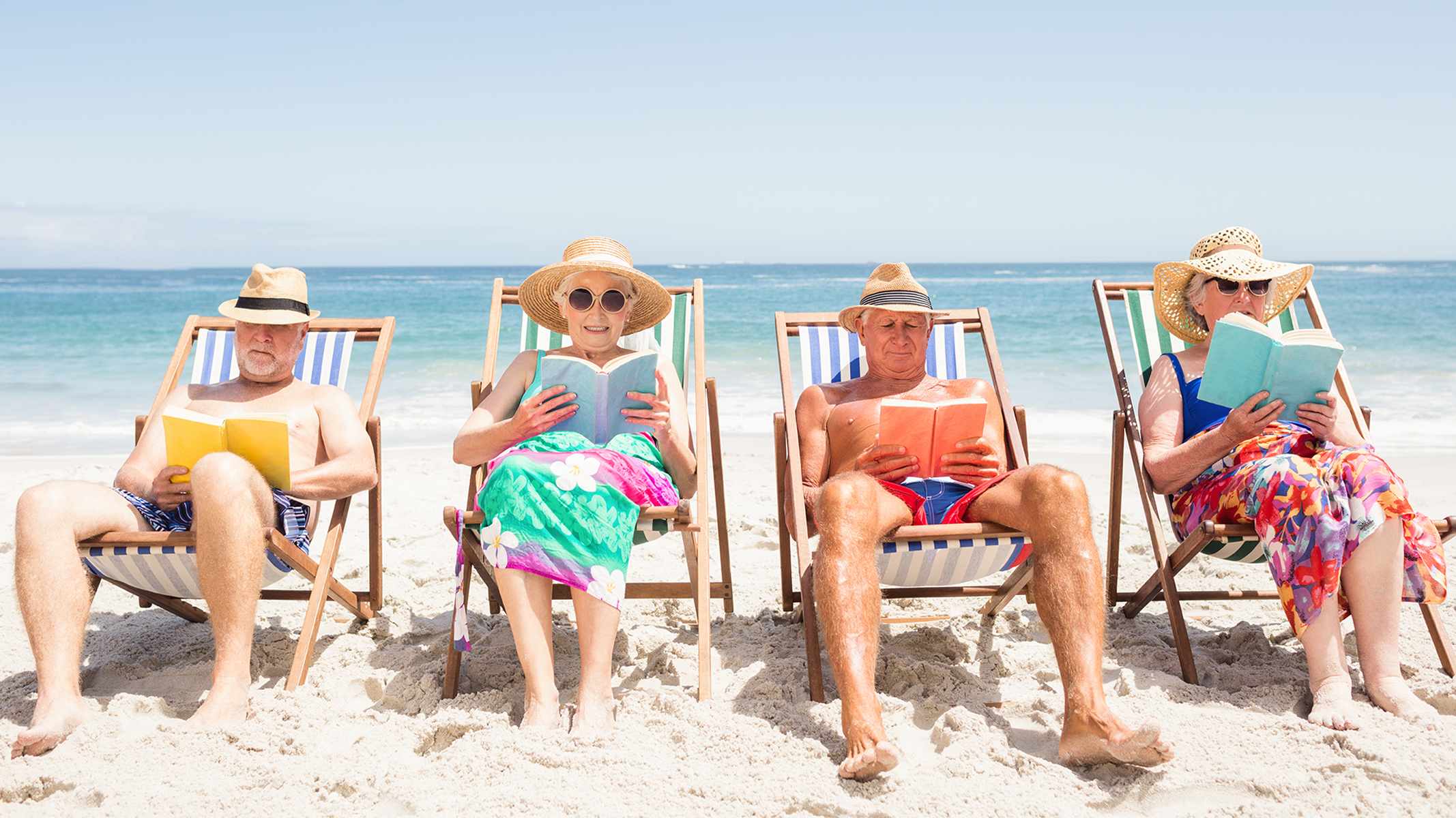 Cuatro personas mayores leyendo libros y descansando en la playa.​​ 